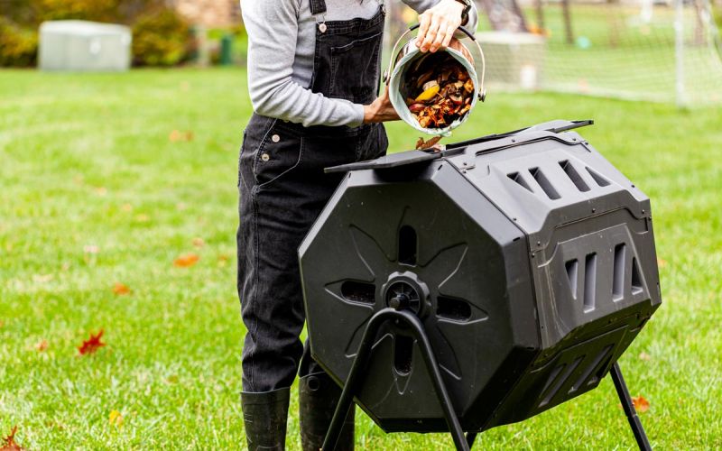 person dumping food scraps from kitchen pail into backyard compost tumbler