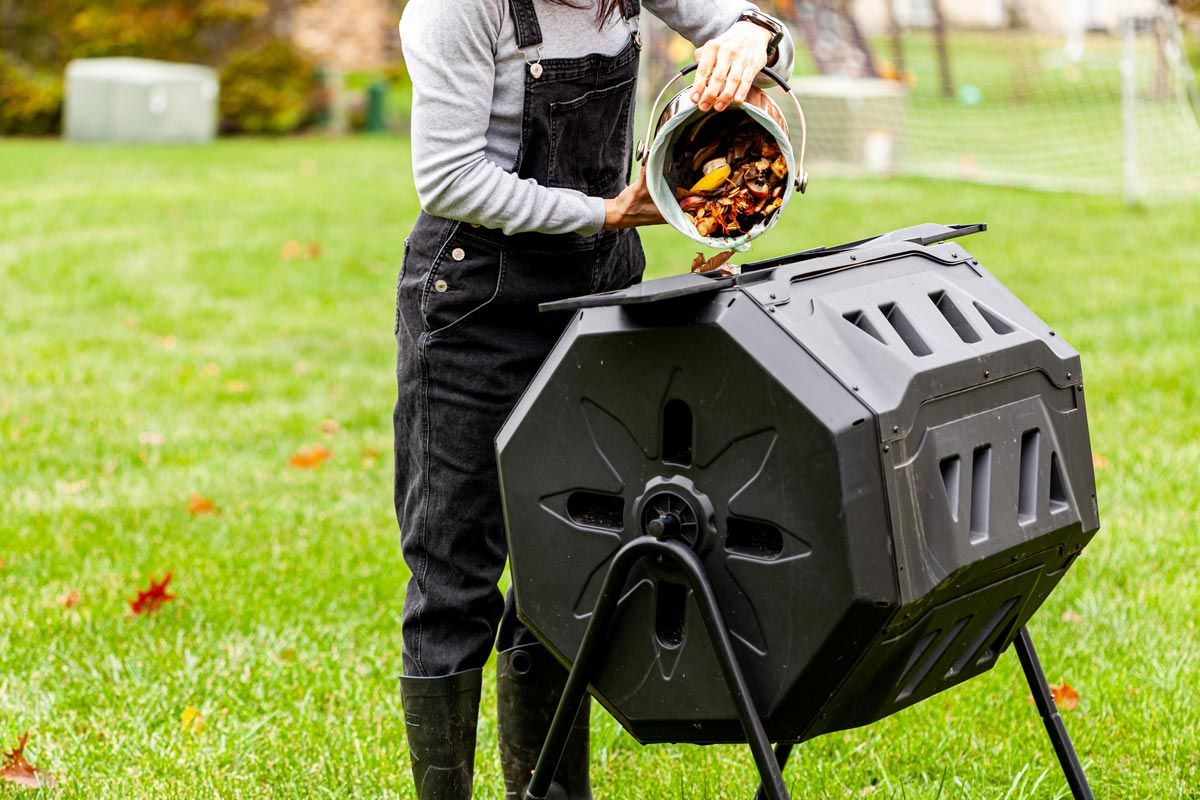 person dumping food scraps from kitchen pail into backyard compost tumbler