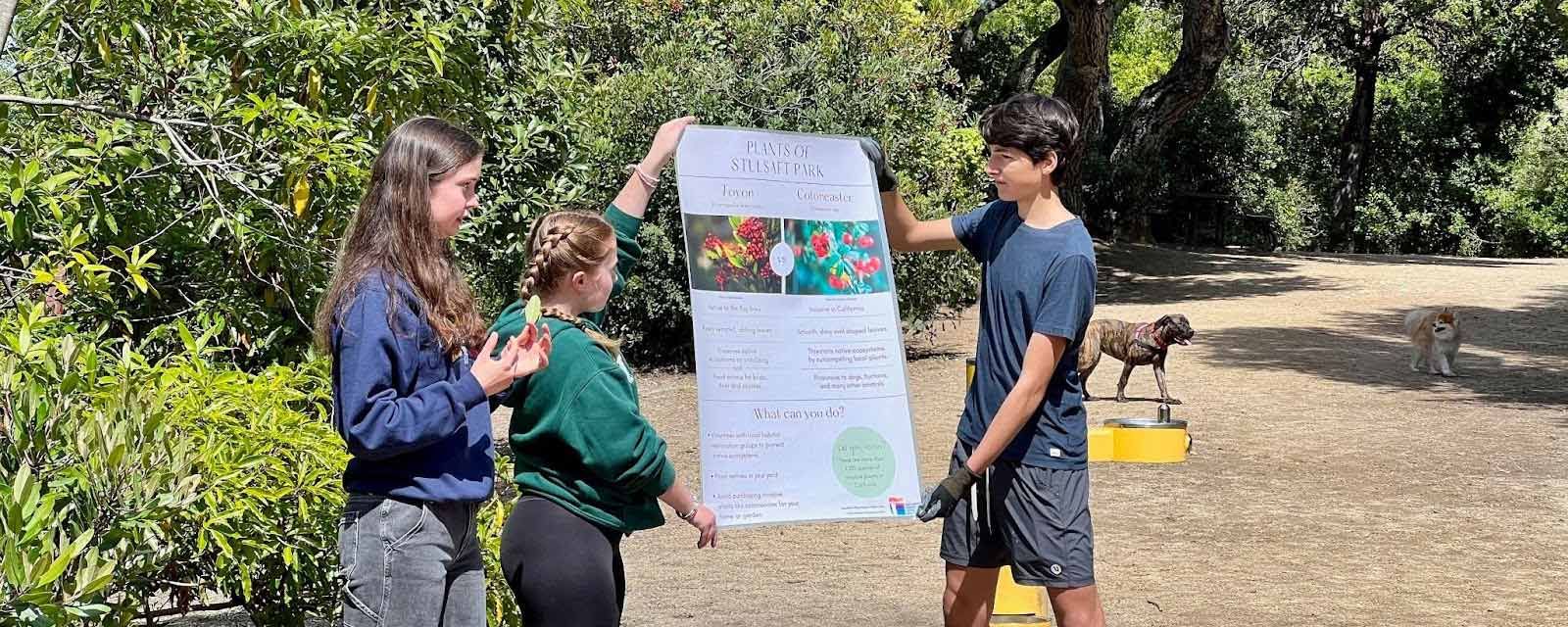 three students in Youth Climate Ambassadors program hold up their project poster in  local park